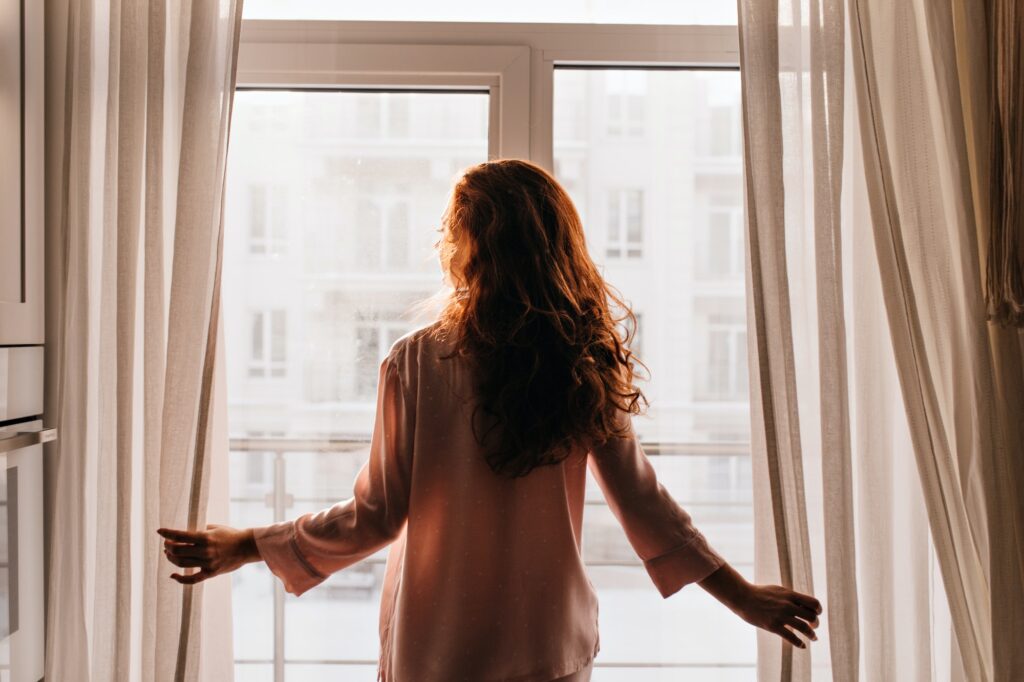 Red-haired girl touching curtains. Indoor photo of caucasian young lady standing near window.